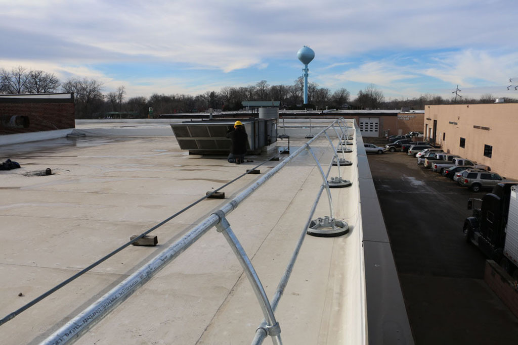 Galvanized architectural guardrail on non-penetrating bases along a roof parapet, with a worker servicing a rooftop unit beyond