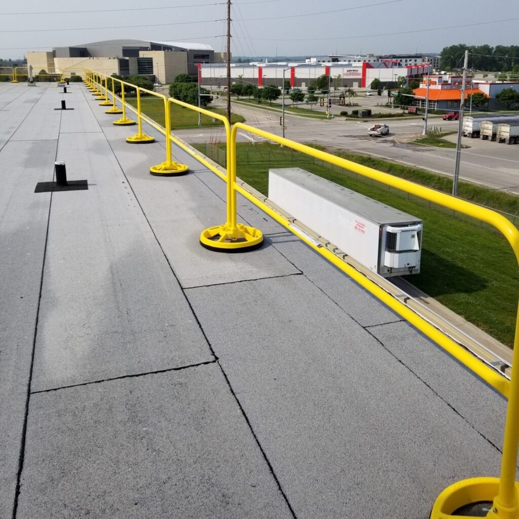A long run of yellow non-penetrating guardrail along a flat roof edge on counterweighted bases